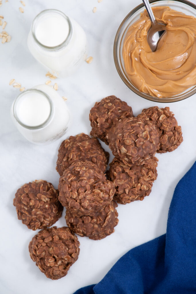 stack of chocolate no bake cookies with two glasses of milk and a bowl of peanut butter